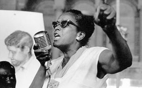 Photograph of human rights activist Ella Baker speaking into a microphone with her fist raised.