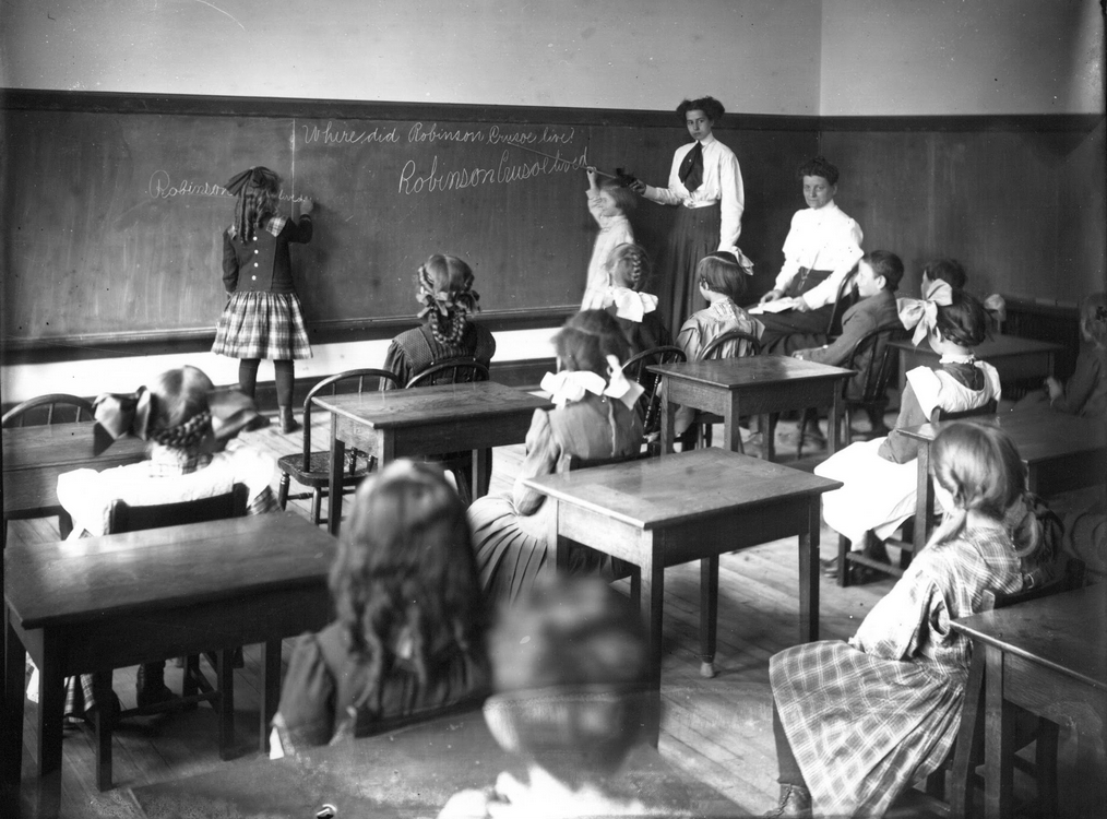 Historical picture of an elementary classroom. Students are all white and sitting in straight rows facing the chalk board.