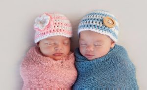 Two very young babies, sleeping side by side. The one on the left is swaddled in a pink blanket and is wearing a pink and white knit cap with a pink and white flower on the side. The one on the left is swaddled in a blue blanket and is wearing a blue and white knit cap with a big button on the side.