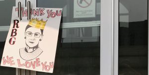 Image shows a handmade sign hung on two door handles. The image shows a drawing of Ruth Bader Ginsburg in a gold crown. The text reads "Thank you RBG. We Love you."